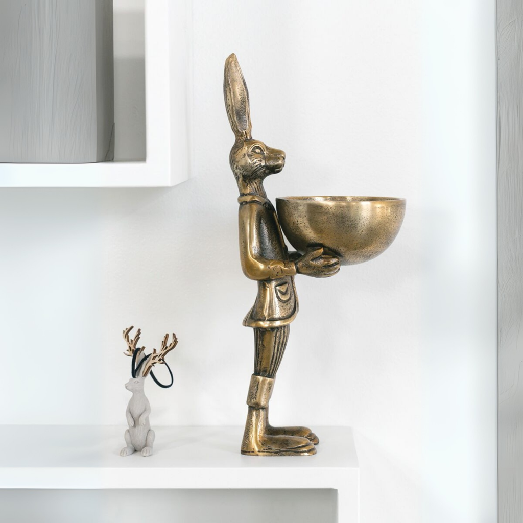 Decorative brass hare figurine holding a bowl, displayed on a modern white shelf with a small deer ornament nearby