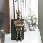 Vintage-style rabbit figurine holding a small bowl, placed on a white surface with candles in the background