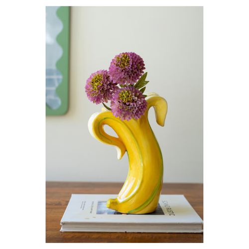 Yellow banana-shaped ceramic vase holding purple flowers on a wooden table with books underneath