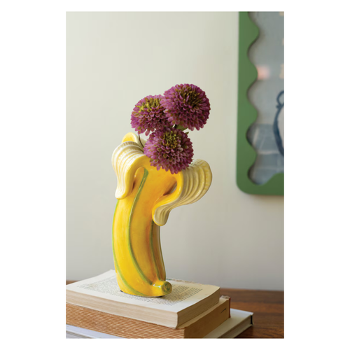 Yellow ceramic banana-shaped vase holding purple flowers on stacked books with a blurred background.