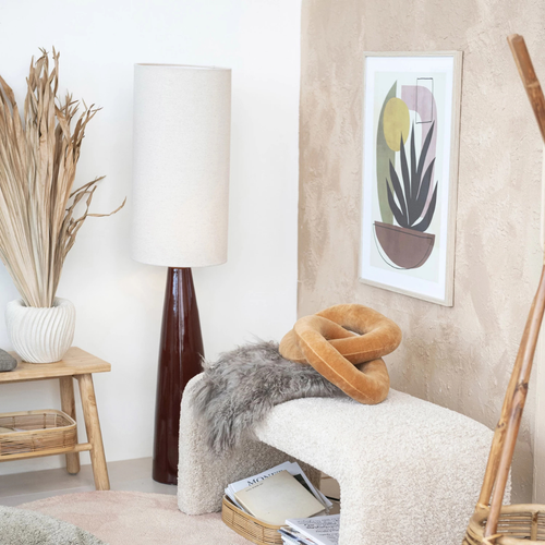 Tall ceramic lamp with white cylindrical shade beside a cozy bench and modern abstract wall art in a soft neutral room.