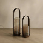Two modern ribbed glass lanterns in amber and smoky brown tones with dark metal arched handles, standing on a beige surface against a neutral beige background.