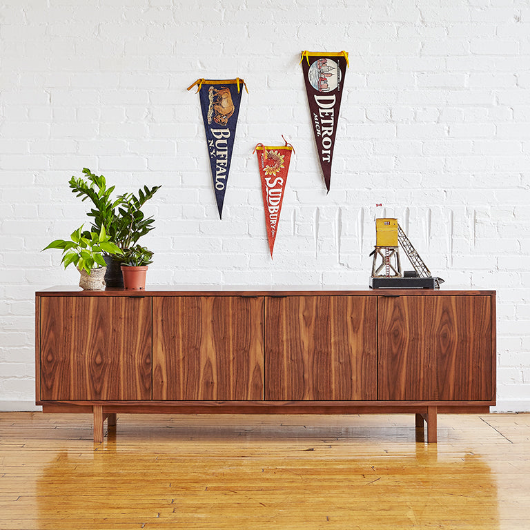 Midcentury modern walnut credenza with plants, decorative objects, and vintage pennants on white brick wall.