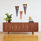 Midcentury modern walnut credenza with plants, decorative objects, and vintage pennants on white brick wall.