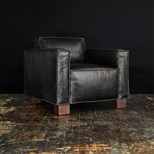 Black leather armchair with wide square arms and wooden block legs on a rustic floor background