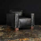 Black leather armchair with wide square arms and wooden block legs on a rustic floor background