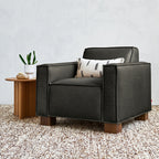 Modern black leather chair with wooden legs and a decorative patterned pillow on a textured rug next to a wooden side table with plants