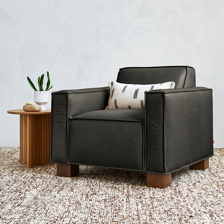 Modern black leather chair with wooden legs and a decorative patterned pillow on a textured rug next to a wooden side table with plants