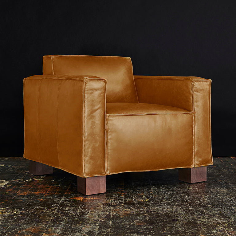 Brown leather armchair with wide square arms and wooden block legs on a textured floor.