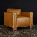Brown leather armchair with wide square arms and wooden block legs on a textured floor.