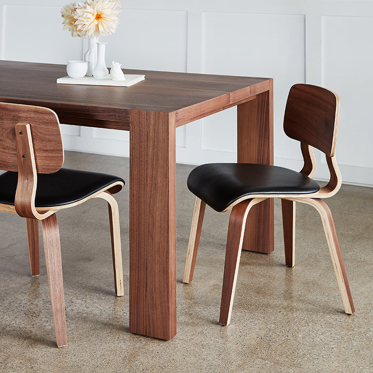 Two modern dining chairs with curved walnut backs and black leather seats next to a matching wood table.