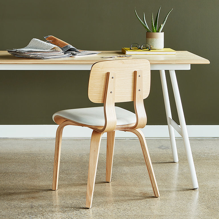 Light wood dining chair with curved backrest and white cushioned seat paired with a minimalist wooden desk and plant decor
