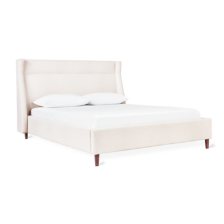 Cream upholstered bed with a tall padded headboard and wooden legs, styled with two white pillows and mattress
