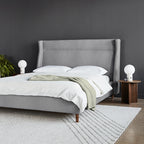 Modern grey upholstered bed with tall wingback headboard, white bedding, wooden legs, bedside table and lamps in bedroom setting