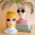 Two colorful ceramic lady vases wearing sunglasses, one with a bow and one with a headband, displayed with books and plant leaves.