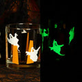 A clear glass tumbler with Halloween ghost and pumpkin cutout designs, shown on a dark patterned surface next to a skull prop, with a split view on the right showing the glass glowing green in the dark.