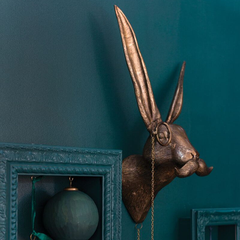 Bronze-colored wall mounted hare sculpture with long ears against a dark teal painted wall in modern decor setting