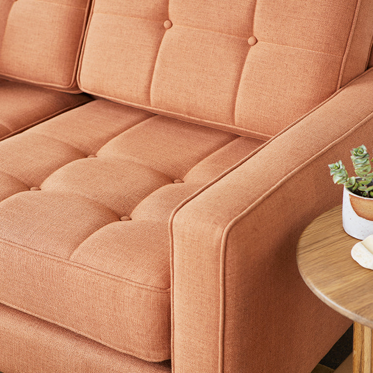 Close-up of a tufted burnt orange fabric sectional sofa armrest next to a wooden side table with a small potted plant