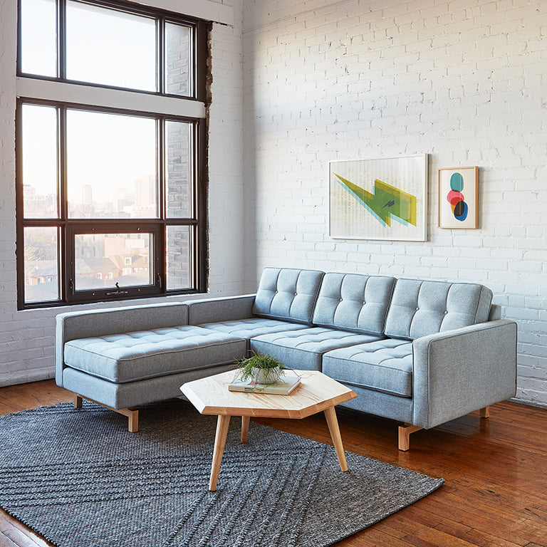 Modern light gray sectional sofa with tufted cushions in a bright room featuring artwork and a geometric coffee table.