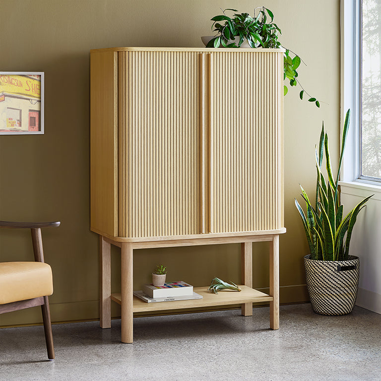 Light oak tall cabinet with vertical slatted doors, slim legs, and decorative plants on and beside it in a modern room.