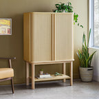 Light oak tall cabinet with vertical slatted doors, slim legs, and decorative plants on and beside it in a modern room.