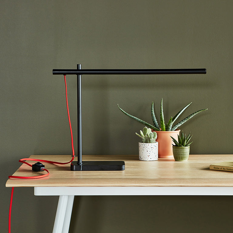 Sleek black task lamp with red cord on wooden desk next to three small potted succulents and a book.