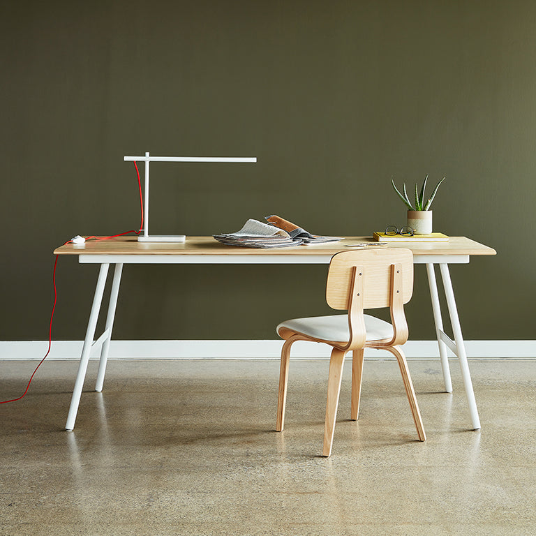 Minimalist task lamp with white frame and red cord on a wooden desk with chair and potted plant against olive wall