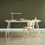 Minimalist task lamp with white frame and red cord on a wooden desk with chair and potted plant against olive wall