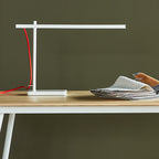 Minimalist white task lamp with a red cord on a wooden desk next to a stack of fabric samples and leather swatches