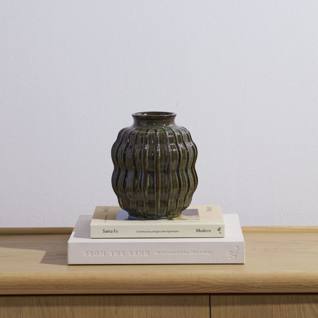 A ribbed ceramic vase in a dark olive green glaze, displayed on top of two stacked books on a light wood surface against a plain white wall.