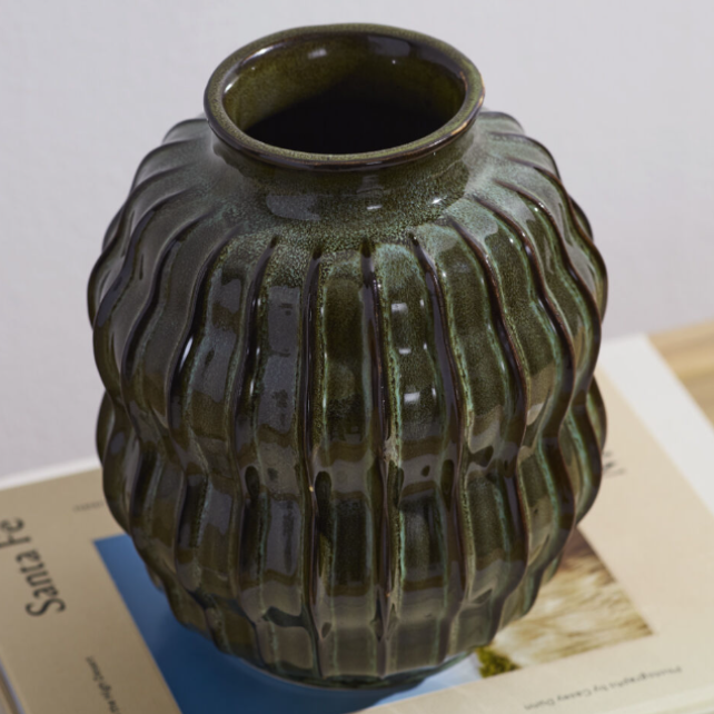 A ribbed ceramic vase with a glossy dark olive green glaze, resting on a book page against a light gray background.