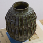 A ribbed ceramic vase with a glossy dark olive green glaze, resting on a book page against a light gray background.