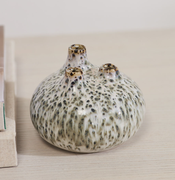 A small ceramic vase with a rounded, bulbous shape and three short necks, covered in a glossy off-white glaze speckled with dark gray and olive-green mottling. It is photographed on a light beige wooden surface next to a neutral-toned book or box.