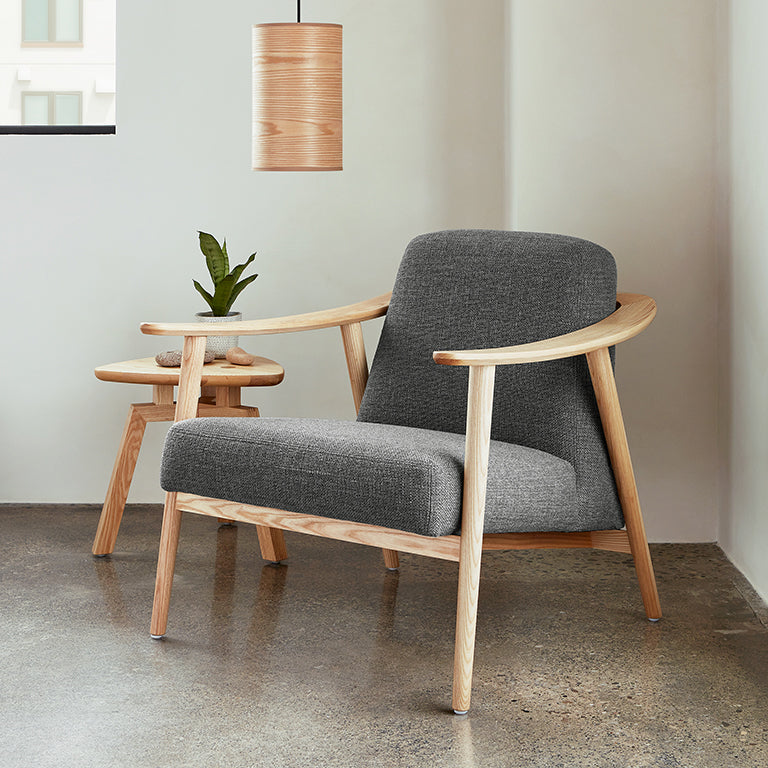 Modern midcentury style armchair with gray cushions and light wood frame next to a small wooden side table and pendant light