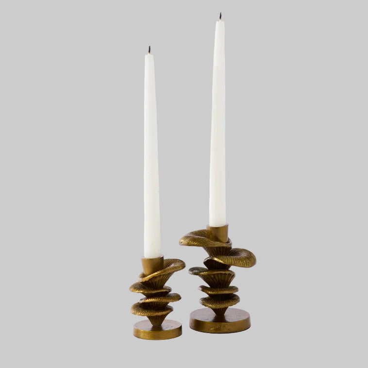 Two sculptural brass candleholders with white tapered candles on a neutral background