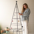 A woman wearing a light blue denim shirt and white pants decorates a black metal spiral Christmas tree with hanging ornaments, set against a white shiplap wall with wrapped gifts on the floor.
