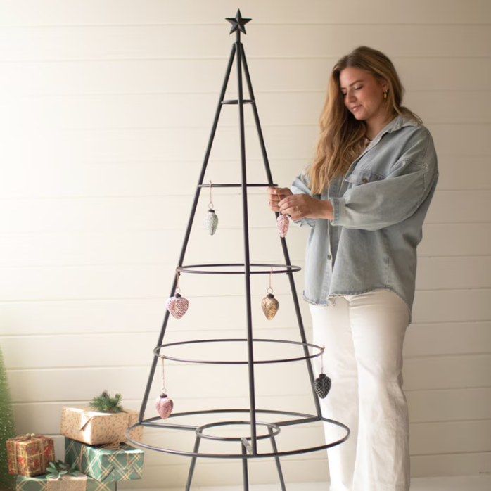 A woman wearing a light blue denim shirt and white pants decorates a black metal spiral Christmas tree with hanging ornaments, set against a white shiplap wall with wrapped gifts on the floor.