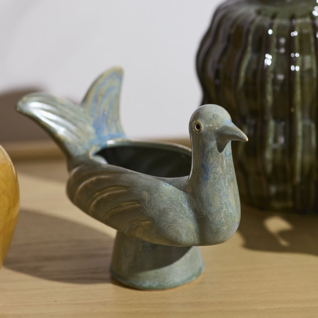 A ceramic bird-shaped planter shaped like a pigeon, featuring a textured blue-gray glaze with subtle light blue accents, set on a light wooden surface.