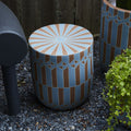 Outdoor ceramic stool with geometric blue and beige pattern on a gravel surface near plants and furniture