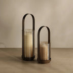 Two ribbed glass candle holders with dark metal handles, one tall and one short, filled with cream-colored candles, set against a warm beige background.