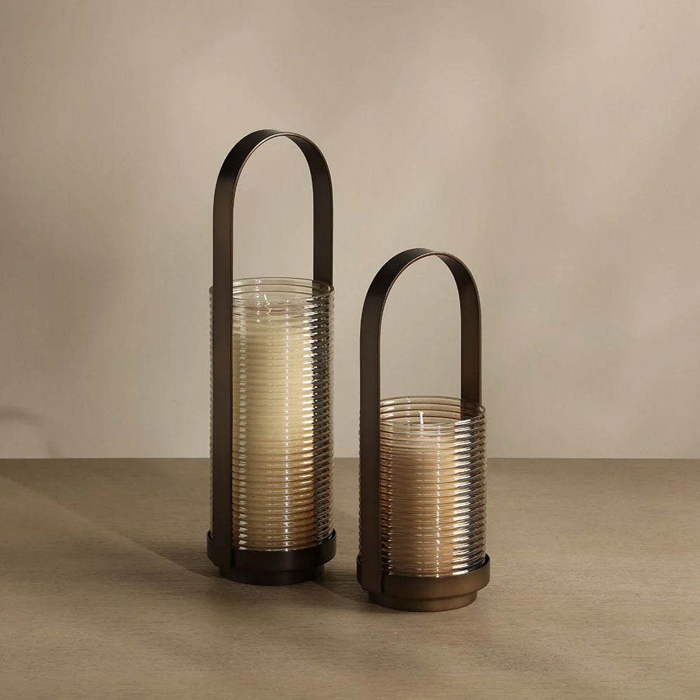 Two ribbed glass candle holders with dark metal handles, one tall and one short, filled with cream-colored candles, set against a warm beige background.
