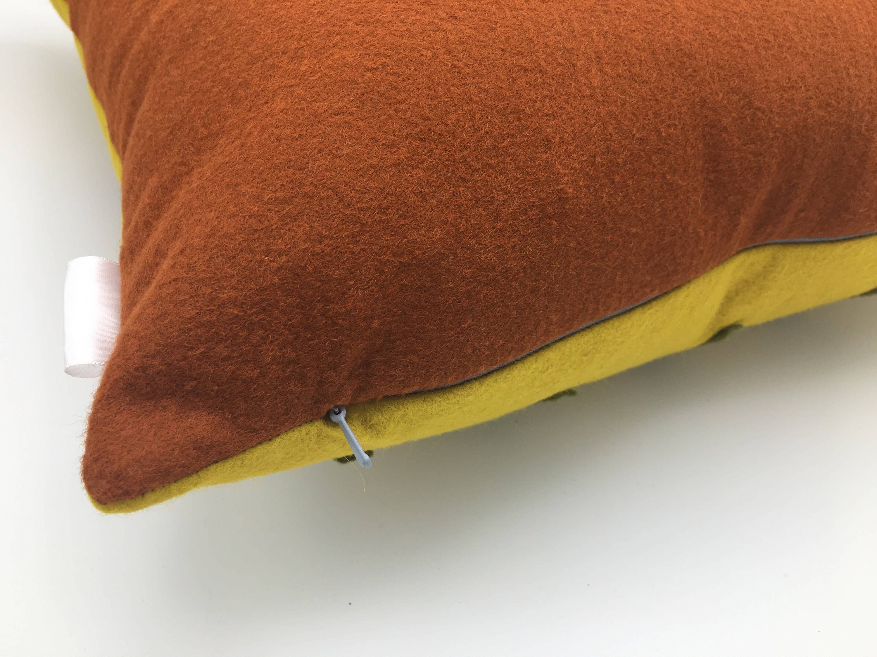 Close-up of a soft wool felt pillow in rust and mustard yellow with a hidden zipper detail on a white background