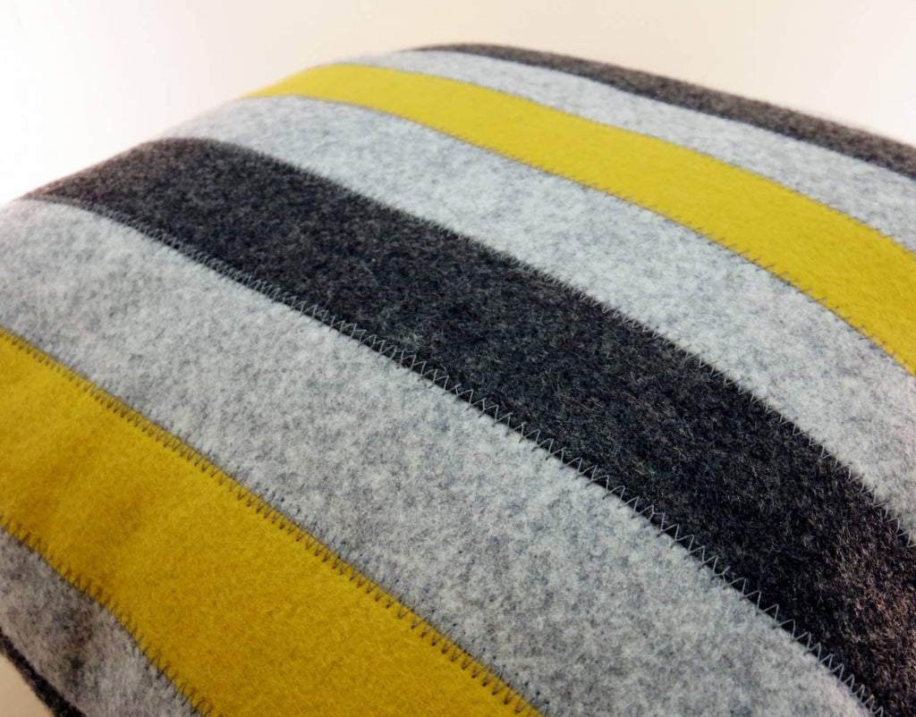 Close-up of a wool felt pillow with alternating yellow, gray, and black stripes and visible stitch detailing.