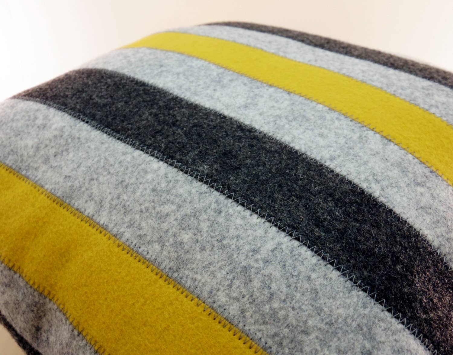 Close-up of a wool felt pillow with alternating yellow, gray, and black stripes and visible stitch detailing.