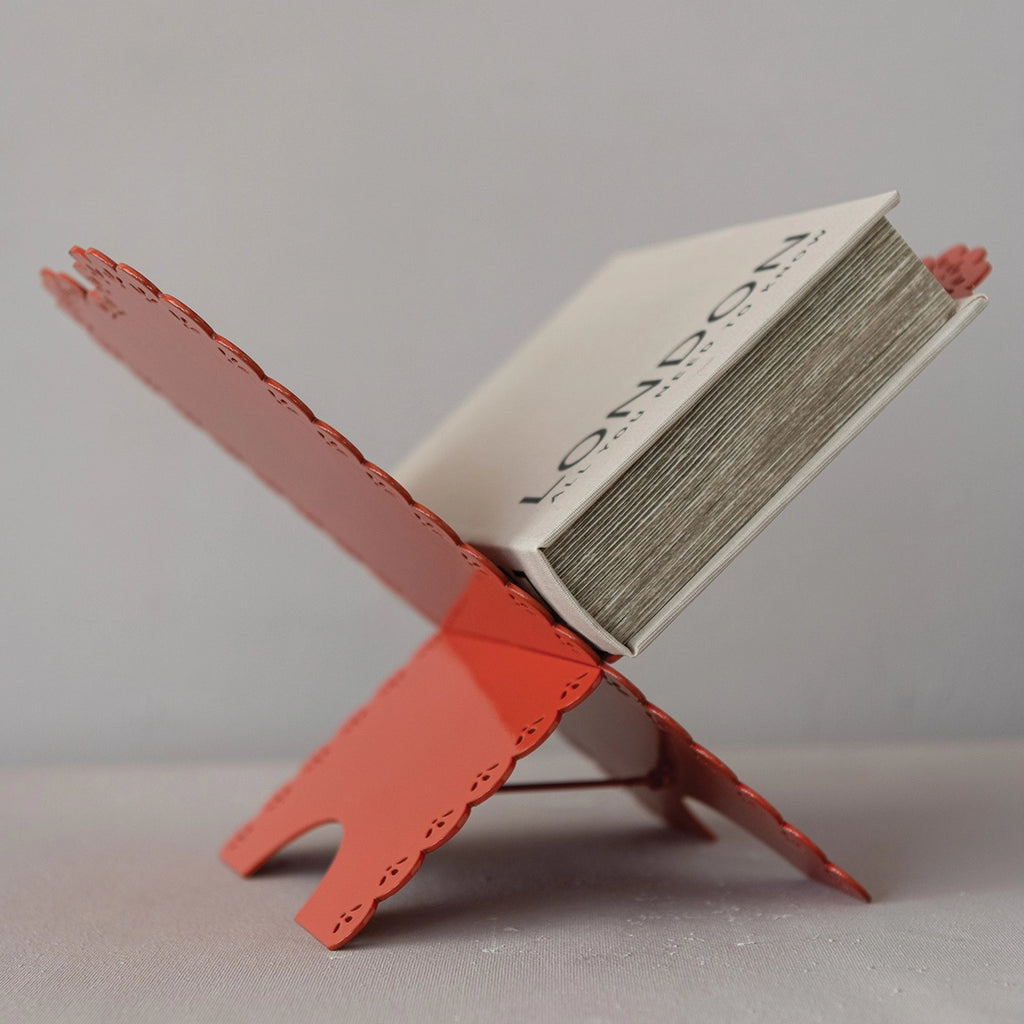 A red metal book stand with scalloped edges holding a book, photographed on a light gray surface against a neutral gray background.