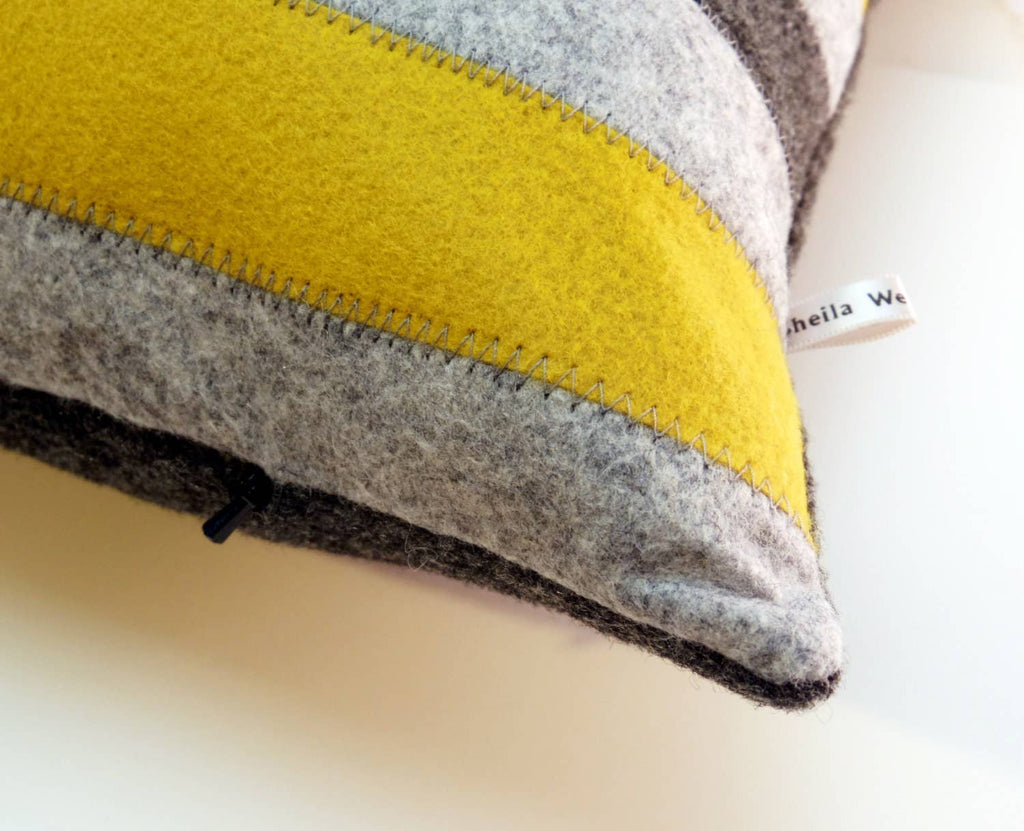 Close-up of a wool felt striped pillow detail showing yellow and gray fabric with visible stitching and a small tag label.