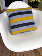 Striped wool felt pillow in gray, black, and mustard yellow resting on a modern white chair outdoors.
