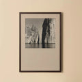 A framed black and white landscape photograph of coastal rock formations over water, mounted on a light beige wall.