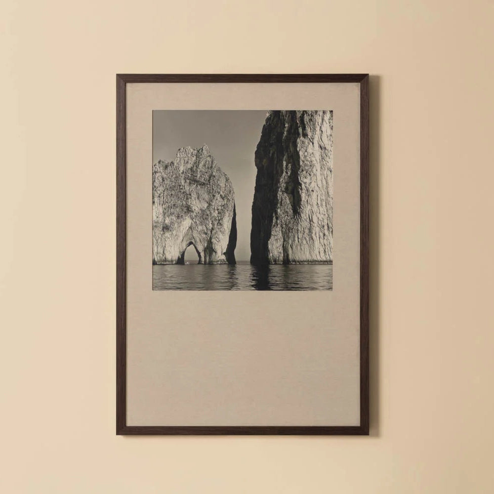 A framed black and white landscape photograph of coastal rock formations over water, mounted on a light beige wall.