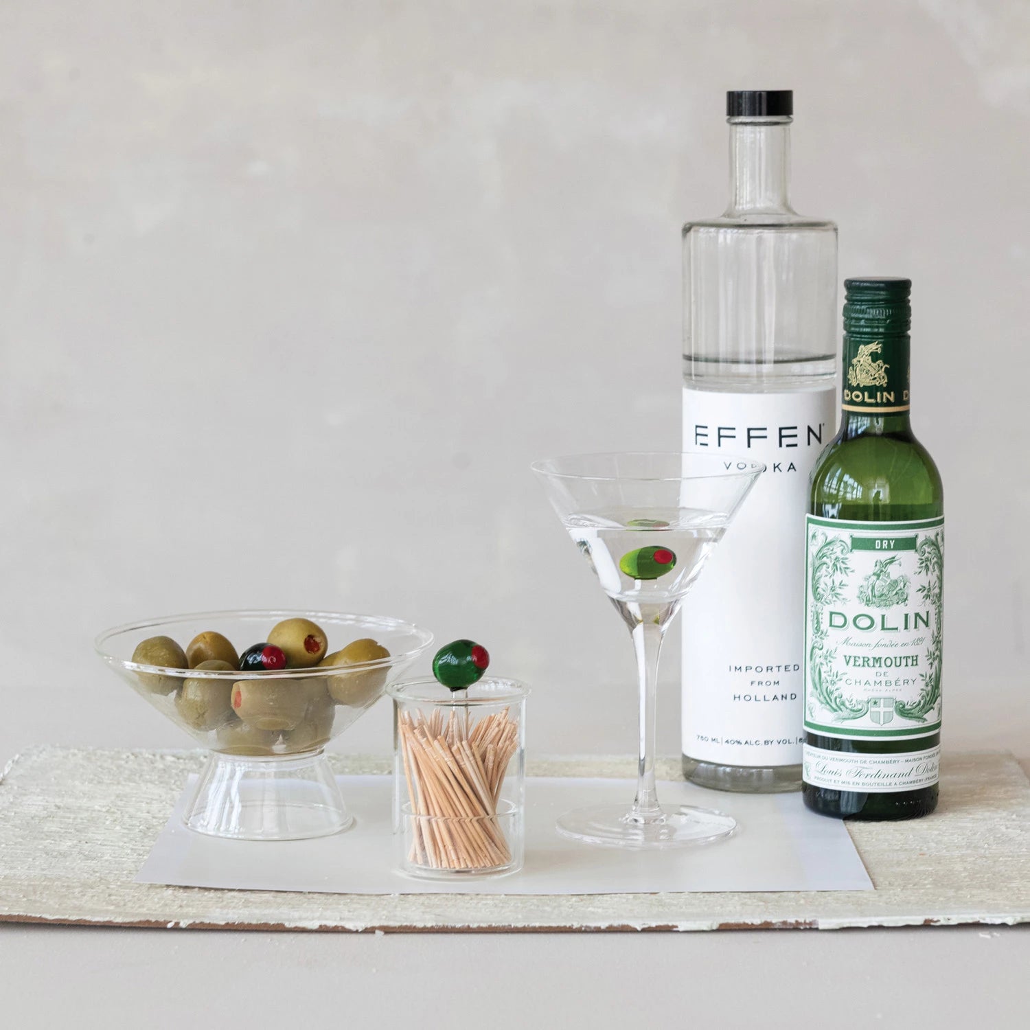 Clear glass olive holder with toothpicks, olives, martini glass, vodka bottle, and vermouth bottle on table.